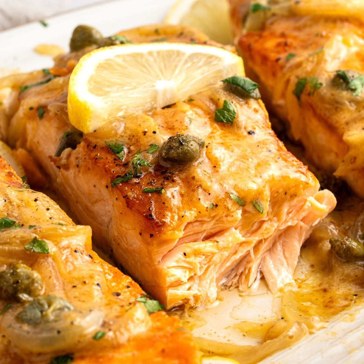 Cooked salmon fillet topped with a lemon slice, capers, and fresh herbs, served with a light golden sauce on a white plate—this elegant salmon piccata brings vibrant flavors to your table.