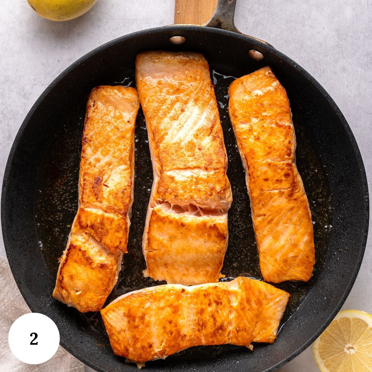 Four golden-brown salmon fillets are cooking in a black skillet, reminiscent of a classic salmon piccata, with a lemon and a partial lemon slice visible nearby. The skillet sits on a light surface. A white circle with the number 2 is in the bottom left corner.