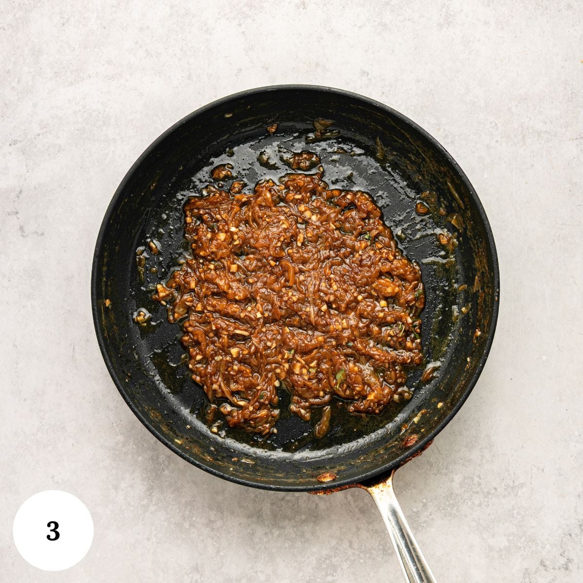 A black skillet with thick, browned, caramelized onions and sauce sits on a light gray surface, hinting at the rich flavors perfect for a greek yogurt french onion dip. A white circle with the number 3 is in the bottom left corner.