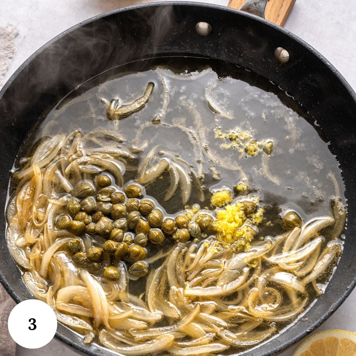 Sliced onions, capers, lemon zest, and sauce simmer in a black skillet for a delicious salmon piccata. Steam rises from the mixture, and a wooden-handled pan sits on a light surface. A lemon wedge is partly visible in the lower right corner.