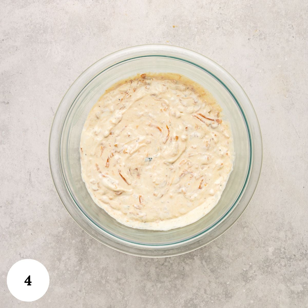 A glass bowl filled with creamy greek yogurt french onion dip containing visible onion pieces sits on a light gray textured surface. A white circle with the number 4 is in the bottom left corner.