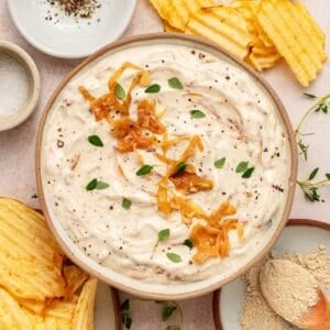 A bowl of creamy greek yogurt french onion dip garnished with caramelized onions and fresh herbs, surrounded by crinkled potato chips and small bowls of seasoning.