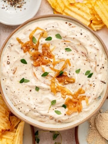A bowl of creamy greek yogurt french onion dip garnished with caramelized onions and fresh herbs, surrounded by crinkled potato chips and small bowls of seasoning.