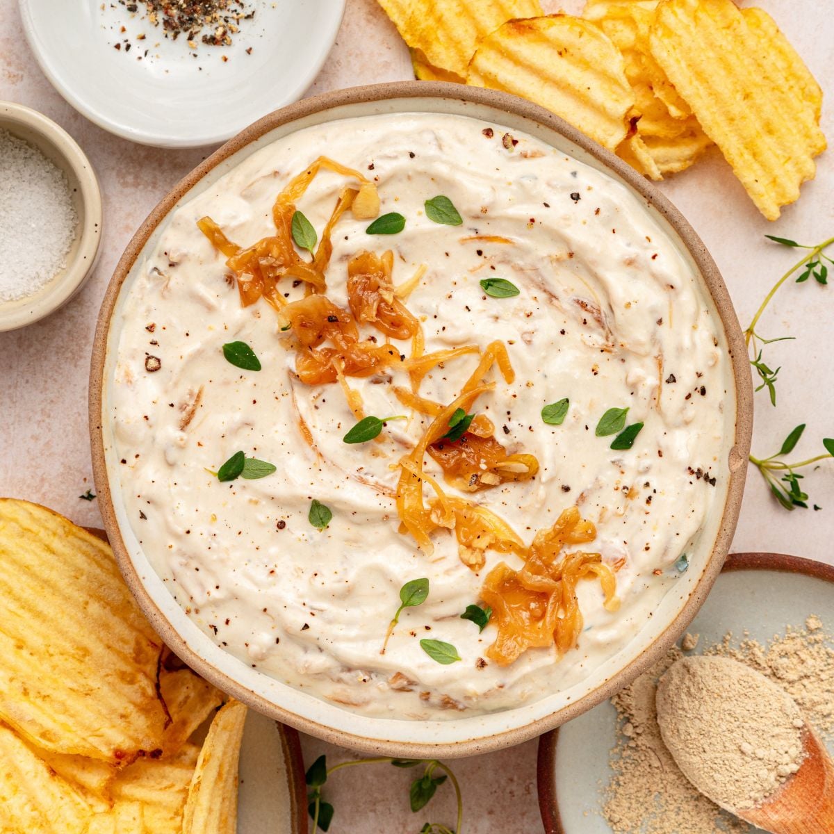 A bowl of creamy greek yogurt french onion dip garnished with caramelized onions and fresh herbs, surrounded by crinkled potato chips and small bowls of seasoning.
