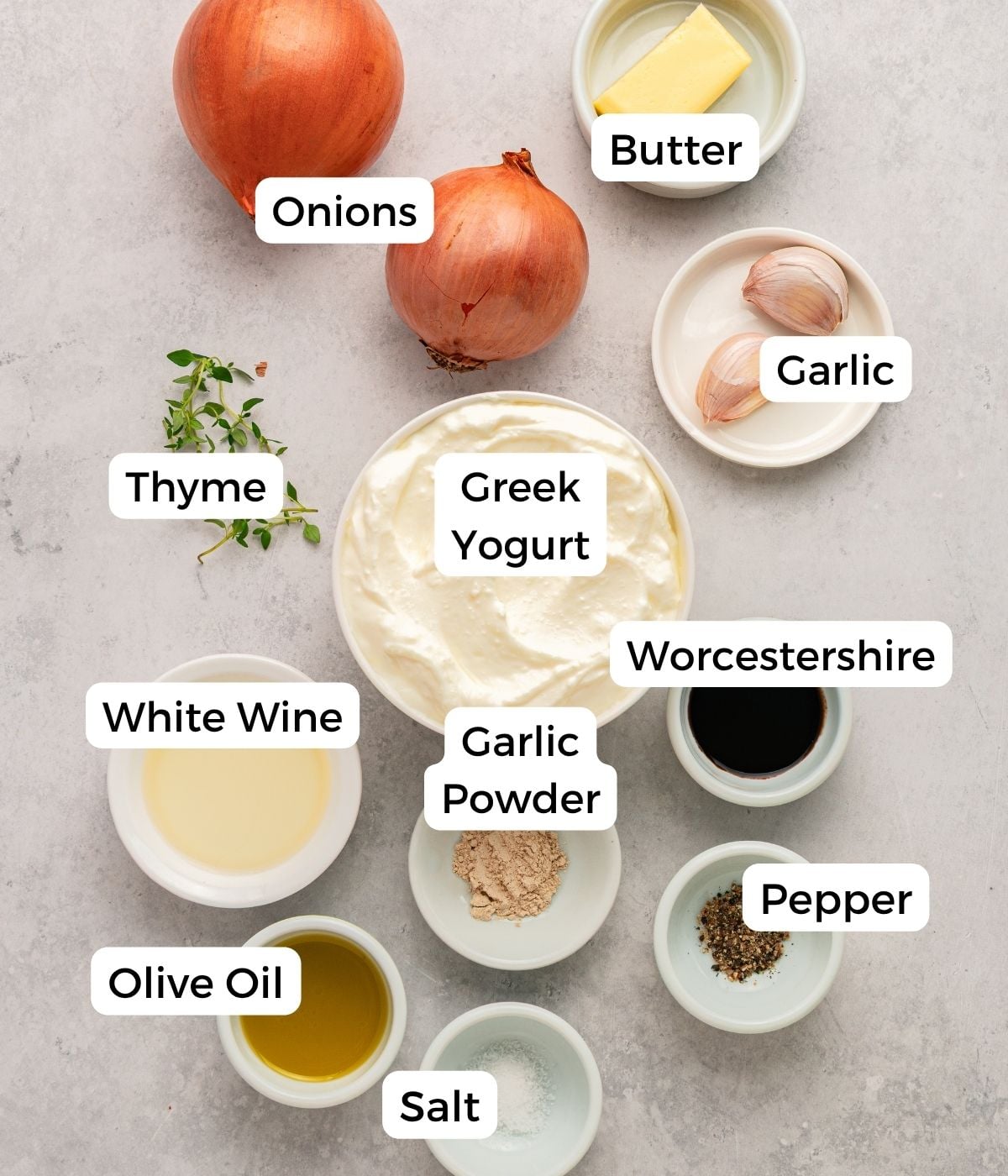Labeled ingredients on a countertop for a rich greek yogurt french onion dip: onions, butter, garlic, thyme, Greek yogurt, garlic powder, Worcestershire sauce, pepper, white wine, olive oil, and salt.