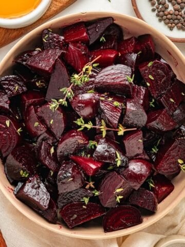 A bowl of roasted beets, cut into pieces and garnished with fresh herbs, is surrounded by small dishes with oil and peppercorns on a light table.