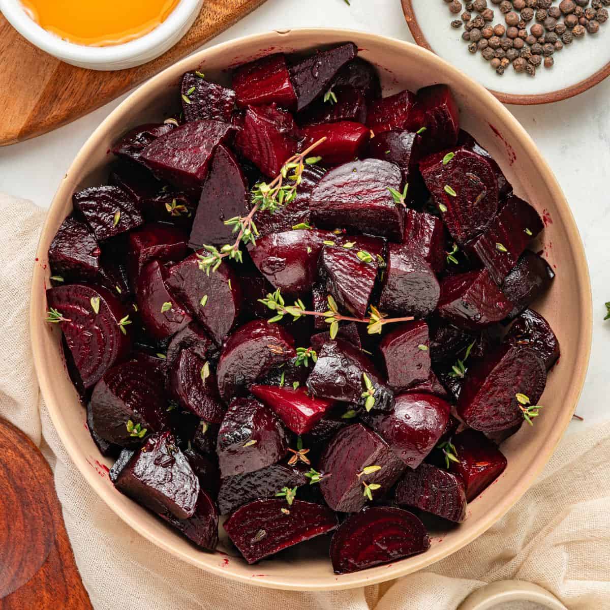 A bowl of roasted beets, cut into pieces and garnished with fresh herbs, is surrounded by small dishes with oil and peppercorns on a light table.