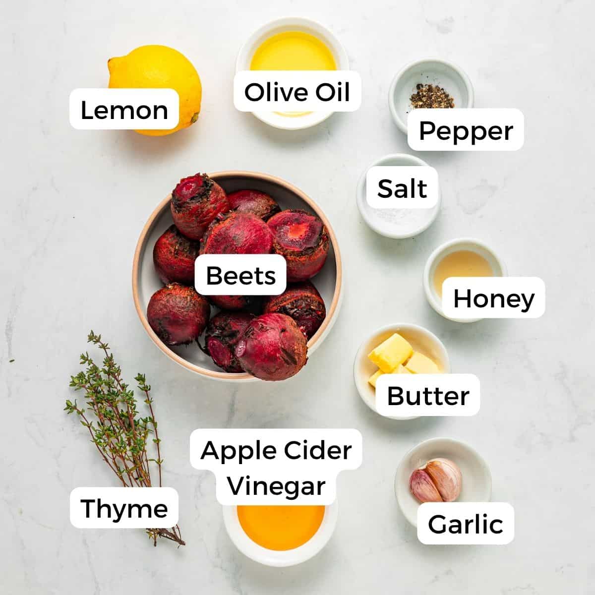 Overhead view of labeled ingredients on a marble surface—lemon, olive oil, pepper, salt, honey, butter, garlic, apple cider vinegar, thyme—surrounding a bowl of roasted beets.