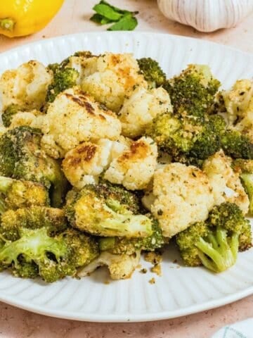 A white plate with roasted broccoli and cauliflower florets, lightly browned, sits on a light surface. Lemons, garlic, and parsley are visible in the background, complementing the vibrant roasted broccoli and cauliflower.