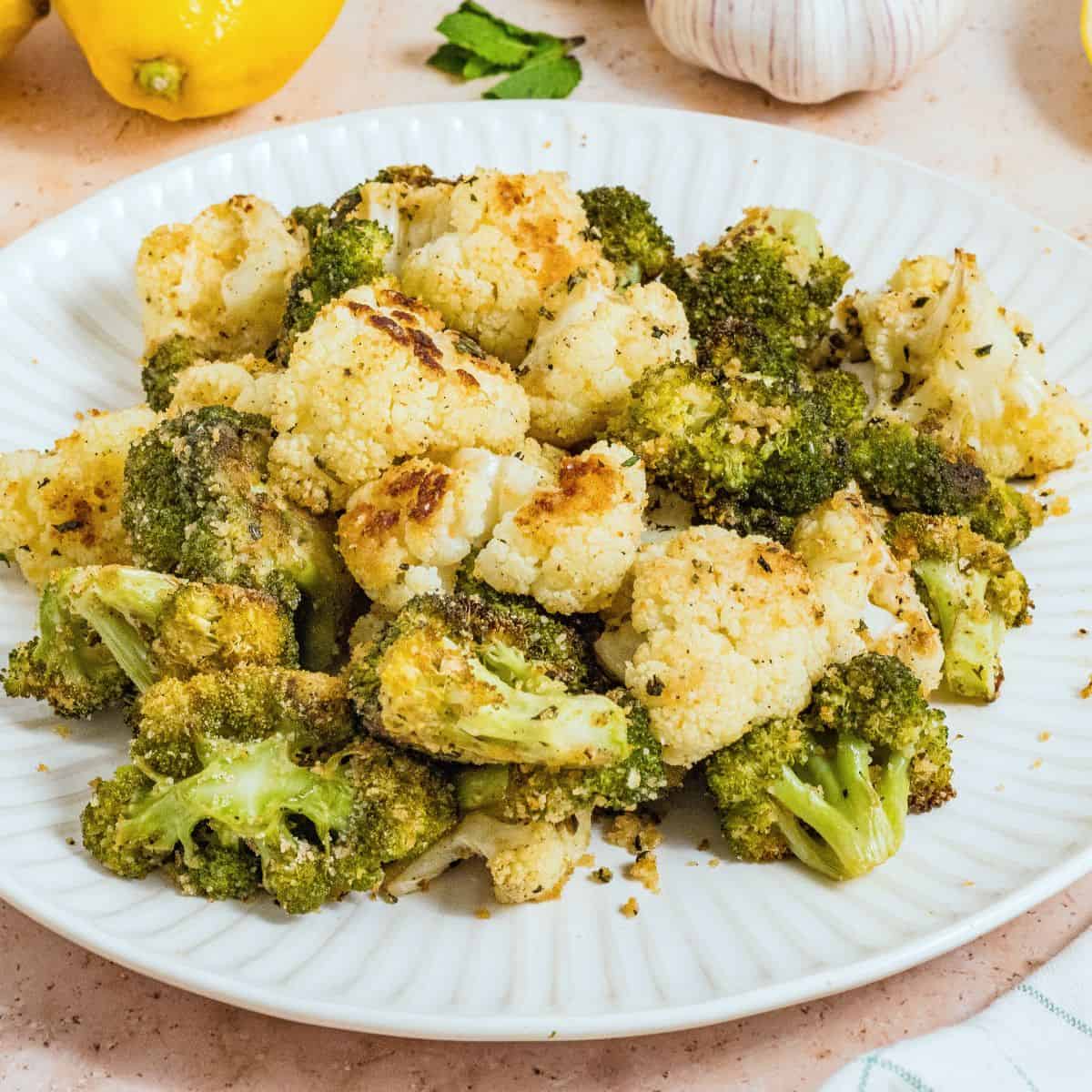 A white plate with roasted broccoli and cauliflower florets, lightly browned, sits on a light surface. Lemons, garlic, and parsley are visible in the background, complementing the vibrant roasted broccoli and cauliflower.