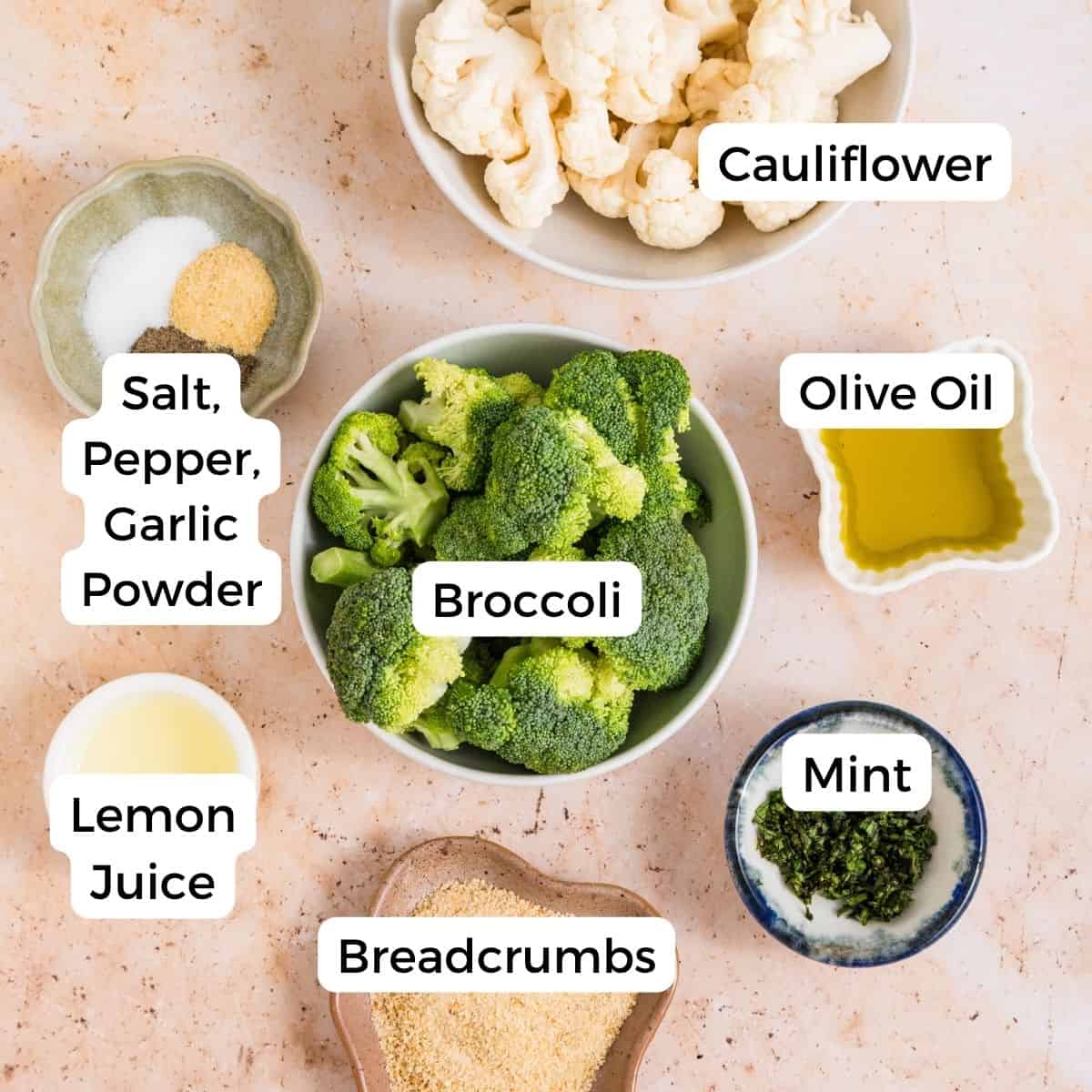 Bowls of ingredients for roasted broccoli and cauliflower—including broccoli, cauliflower, olive oil, breadcrumbs, lemon juice, chopped mint—plus a dish of salt, pepper, and garlic powder—all labeled and arranged on a light-colored surface.