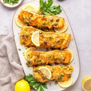 Four baked salmon fillets topped with a creamy sauce and capers, in the style of classic salmon piccata, are garnished with lemon slices and parsley and arranged on an oval white platter. Lemons and chopped herbs are nearby on the light-colored surface.