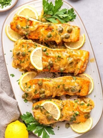 Four baked salmon fillets topped with a creamy sauce and capers, in the style of classic salmon piccata, are garnished with lemon slices and parsley and arranged on an oval white platter. Lemons and chopped herbs are nearby on the light-colored surface.