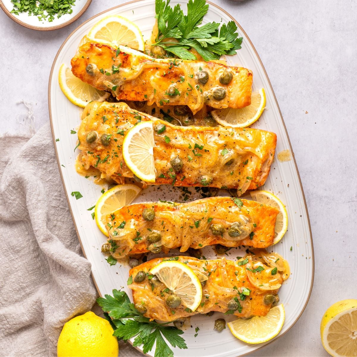 Four baked salmon fillets topped with a creamy sauce and capers, in the style of classic salmon piccata, are garnished with lemon slices and parsley and arranged on an oval white platter. Lemons and chopped herbs are nearby on the light-colored surface.
