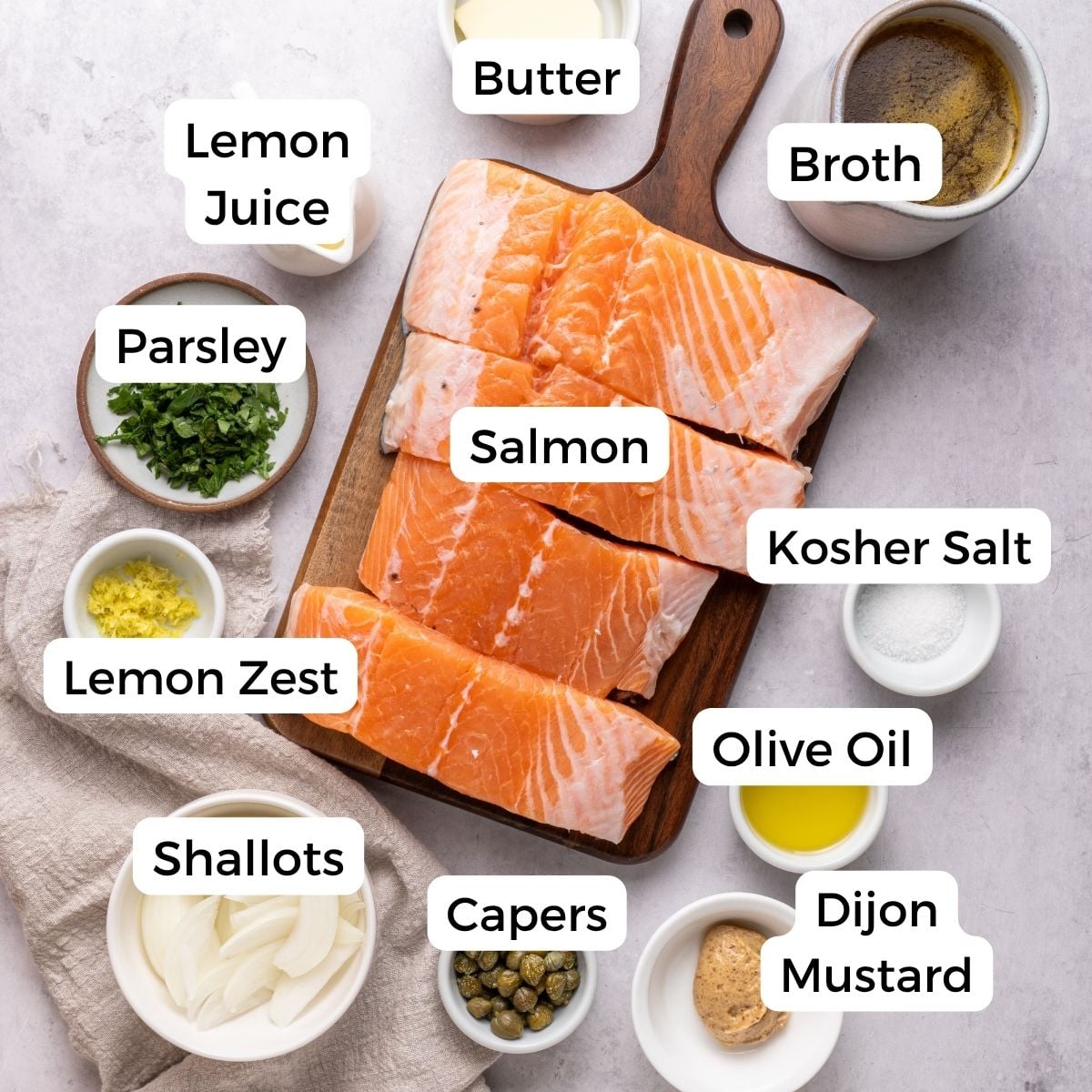Raw salmon fillets on a board surrounded by labeled ingredients—lemon juice, parsley, lemon zest, shallots, capers, olive oil, Dijon mustard, kosher salt, broth, and butter—ready to be transformed into a flavorful salmon piccata.