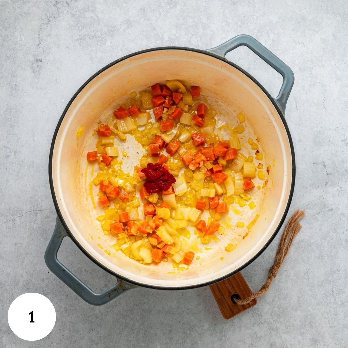 A pot with sautéed chopped carrots, celery, and onion, plus a dollop of tomato paste-classic ingredients for chicken and white bean soup-sits on a light gray surface.