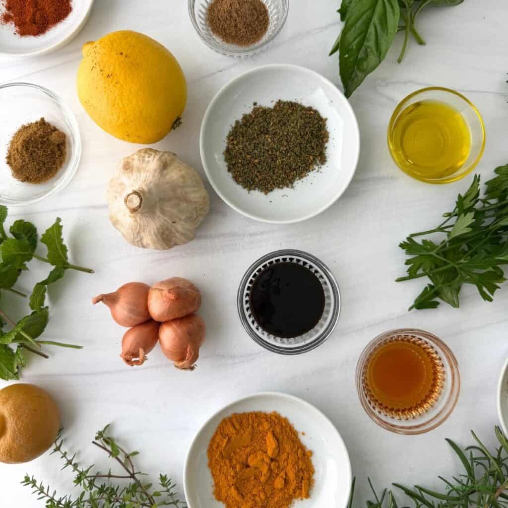 An assortment of herbs, spices, and ingredients including garlic, lemon, shallots, olive oil, vinegar, honey, ground spices, and fresh greens arranged on a white surface-perfect essentials for anyone exploring Mediterranean cooking or looking for a beginner's guide to the vibrant Mediterranean diet.