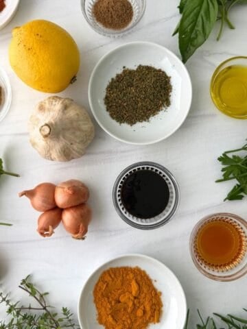 An assortment of herbs, spices, and ingredients including garlic, lemon, shallots, olive oil, vinegar, honey, ground spices, and fresh greens arranged on a white surface-perfect essentials for anyone exploring Mediterranean cooking or looking for a beginner's guide to the vibrant Mediterranean diet.