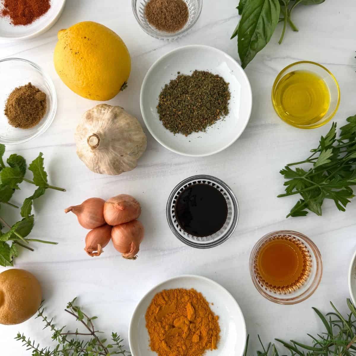 An assortment of herbs, spices, and ingredients including garlic, lemon, shallots, olive oil, vinegar, honey, ground spices, and fresh greens arranged on a white surface—perfect essentials for anyone exploring Mediterranean cooking or looking for a beginner’s guide to the vibrant Mediterranean diet.