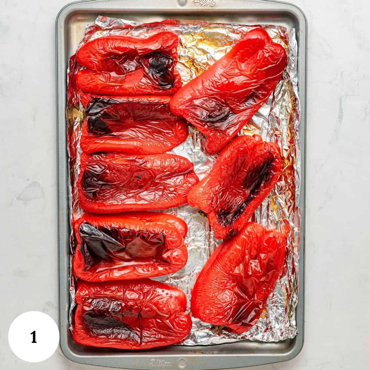 Halved roasted red peppers with charred skin on a foil-lined baking sheet, viewed from above. The roasted red peppers are arranged irregularly and labeled with a number 1 in the bottom left corner.