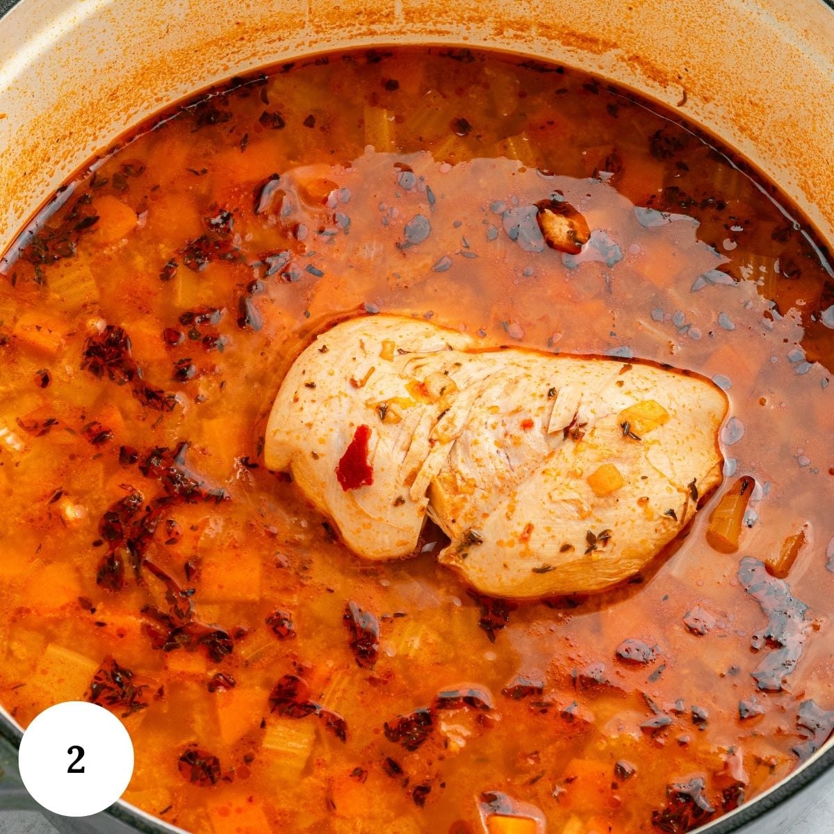 A pot of soup with broth, chopped vegetables, spices, and a large piece of cooked chicken breast in the center evokes all the hearty comfort of a homemade Chicken and White Bean Soup.