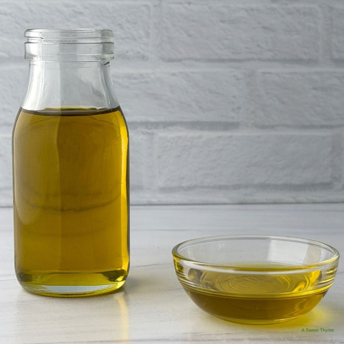A clear glass bottle filled with golden olive oil stands next to a small glass bowl containing olive oil, set on a white countertop with a white brick background—an inviting scene that perfectly captures the essence of Mediterranean cooking.