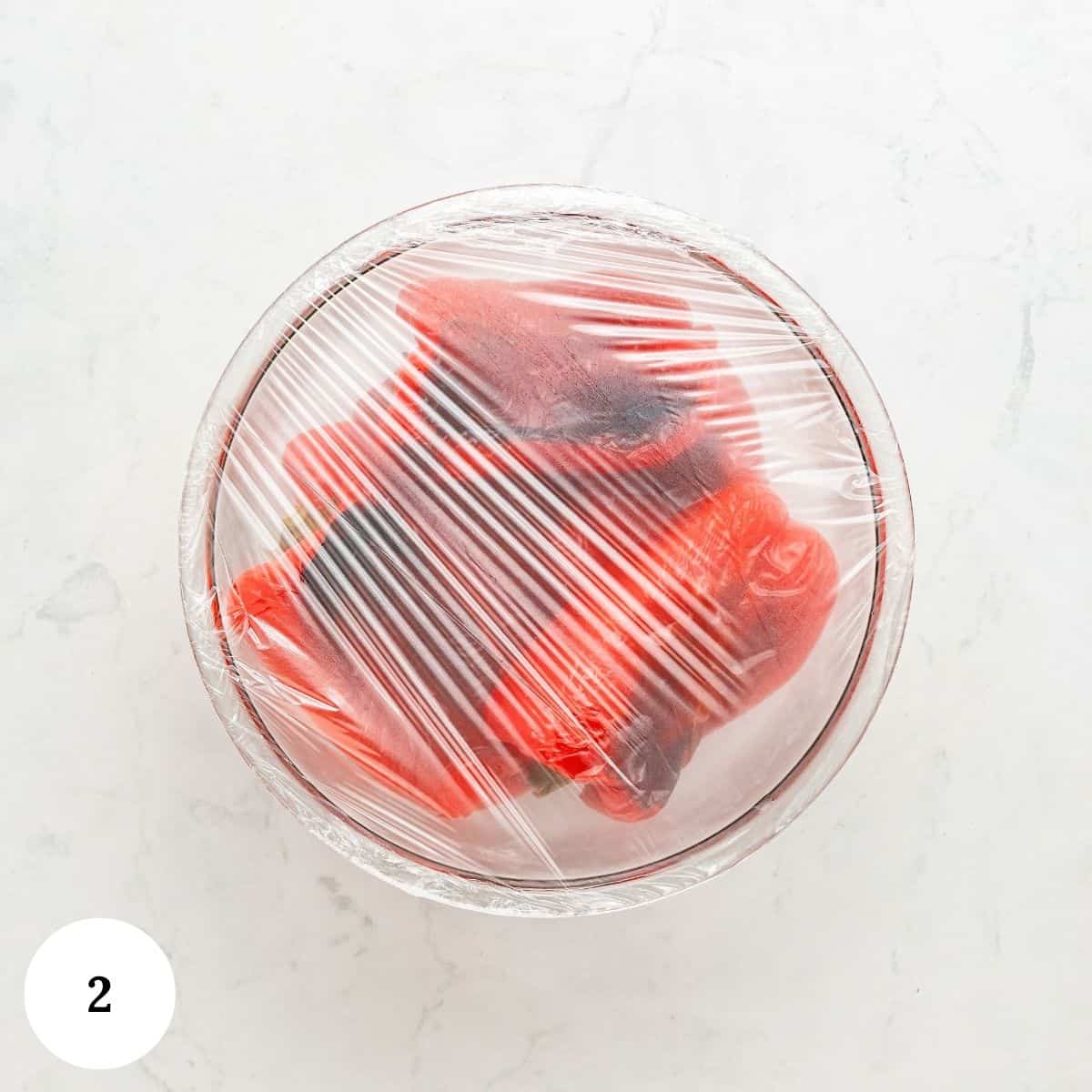 A glass bowl covered with plastic wrap containing roasted red peppers, viewed from above on a white surface. A white circle with the number 2 is in the lower left corner.