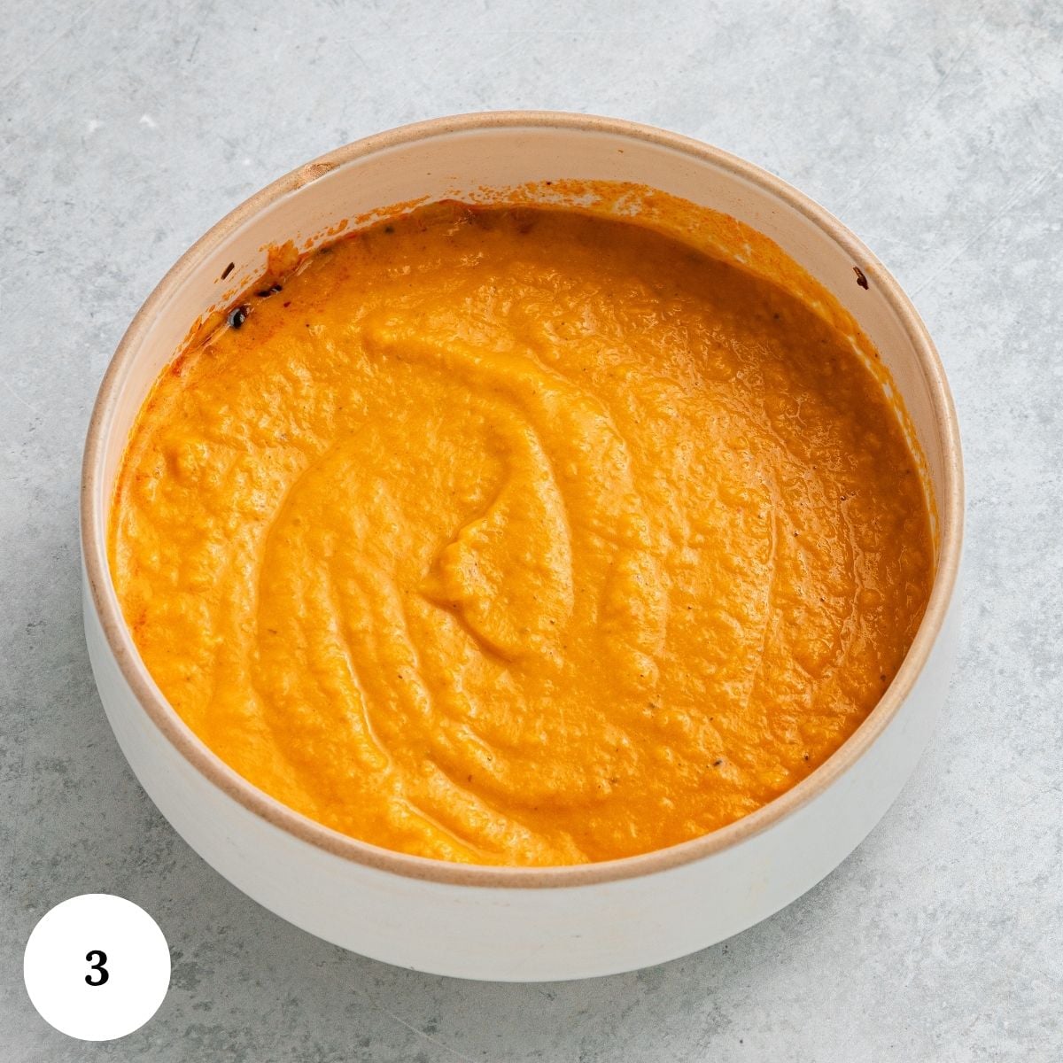 A white bowl filled with smooth orange puree sits on a light gray surface, reminiscent of the comforting hues found in Chicken and White Bean Soup. The number 3 is shown in a white circle in the bottom left corner of the image.