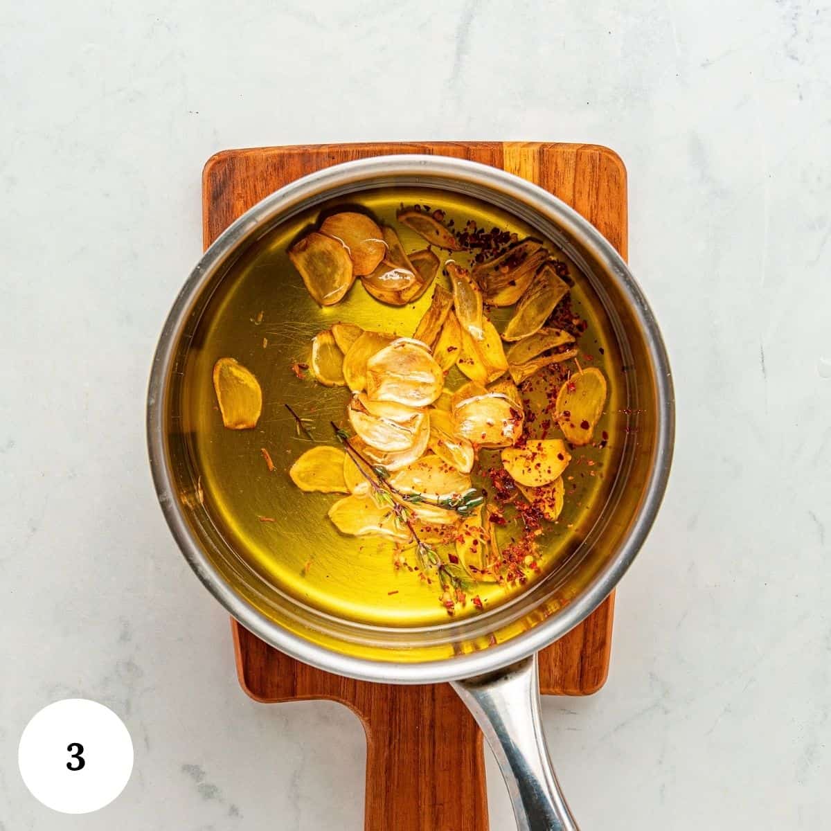 A saucepan of olive oil with sliced garlic, herbs, roasted red peppers, and chili flakes sits on a small wooden board, ready for infusion. A white circle with the number 3 is in the bottom left corner.