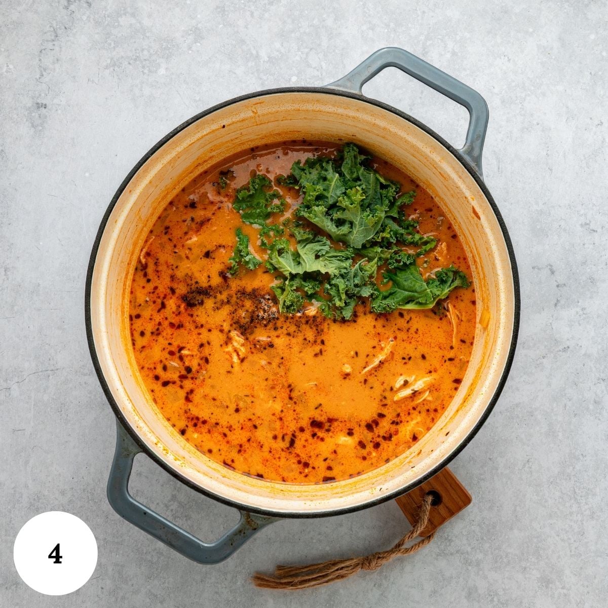 A pot of creamy orange soup garnished with fresh green kale sits on a gray surface. The soup has visible flecks of spices and shredded cheese, reminiscent of hearty Chicken and White Bean Soup, and a wooden-handled spoon rests next to the pot.