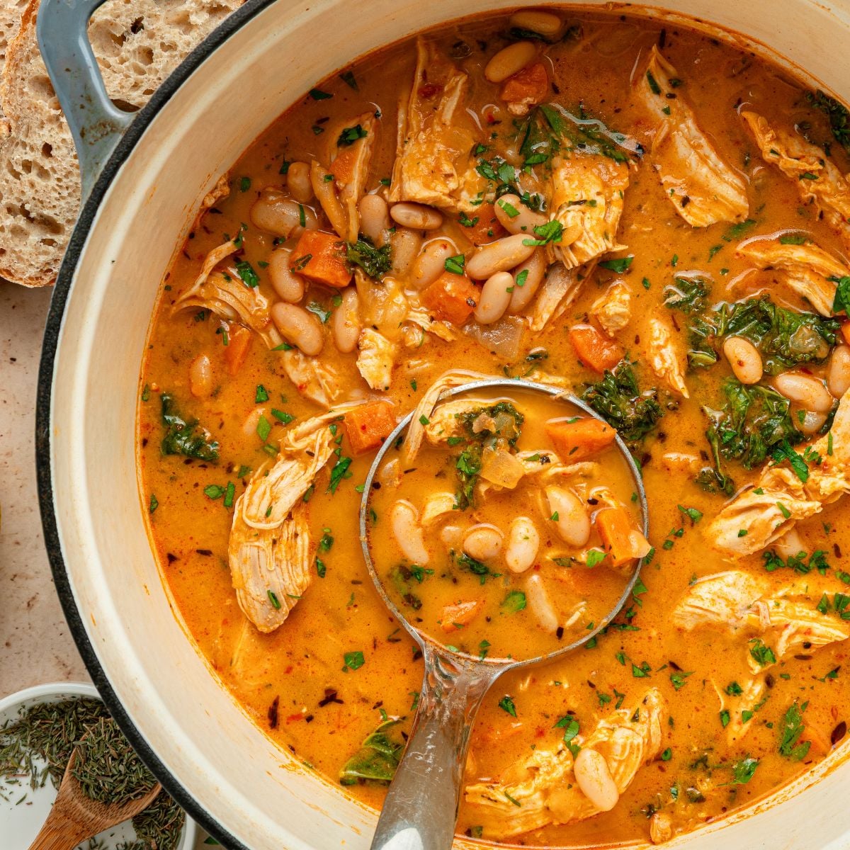 A pot of hearty chicken and white bean soup with greens and chopped vegetables, topped with herbs. A ladle is scooping some Shredded Chicken Soup, and slices of bread are visible on the side.