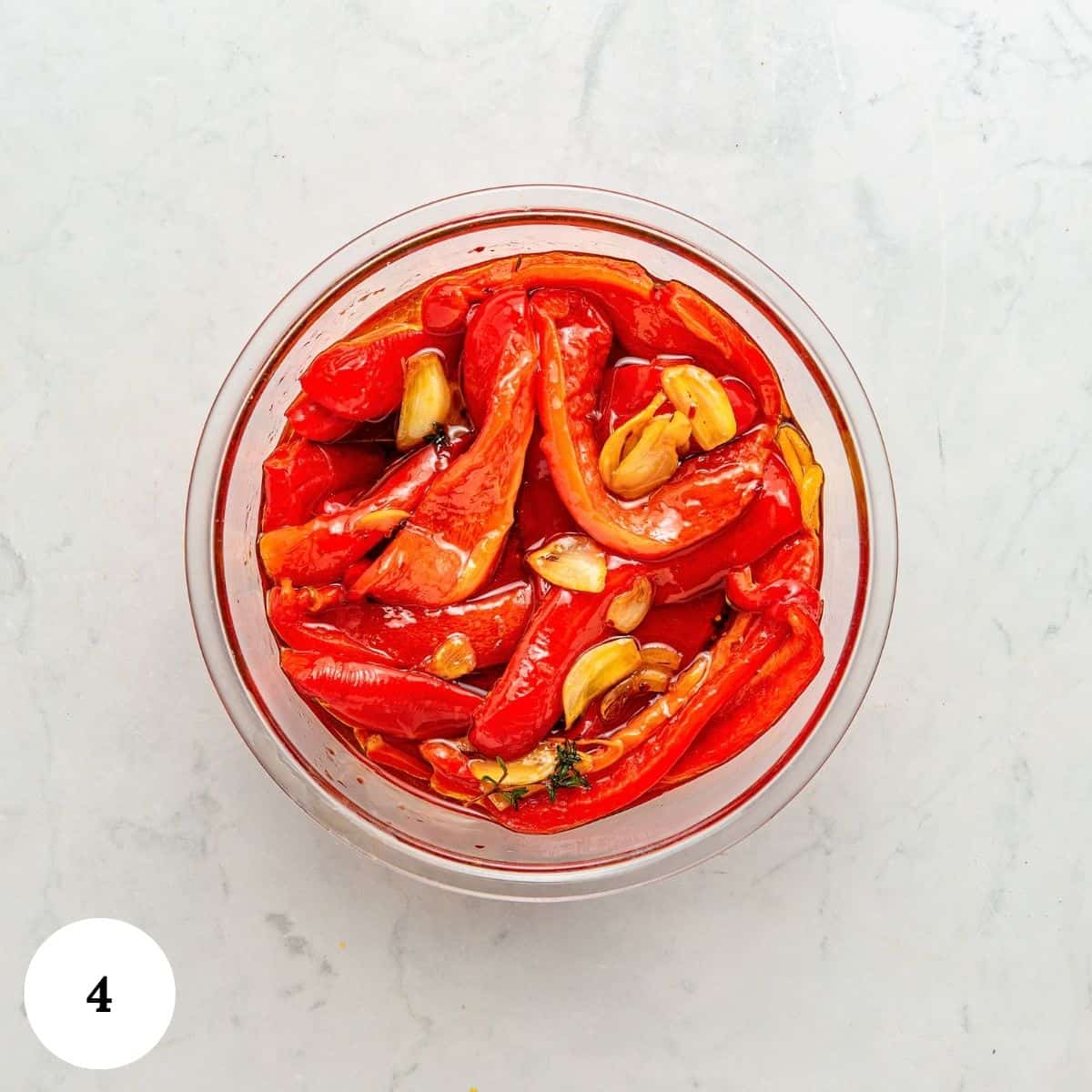 A glass bowl filled with roasted red peppers, garlic cloves, and herbs in olive oil sits on a white marble surface. A white circle with the number 4 is in the lower left corner.