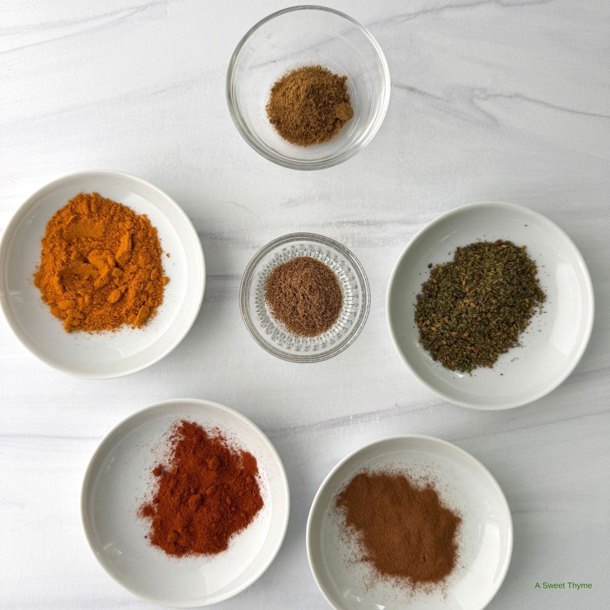 Six small bowls hold different spices, including ground cinnamon, paprika, turmeric, dried herbs, nutmeg, and a brown powder, all arranged in a neat pattern on a white surface—an inviting snapshot of essential flavors often used in Mediterranean cooking. Perfect for those seeking a beginner’s guide to the Mediterranean diet.