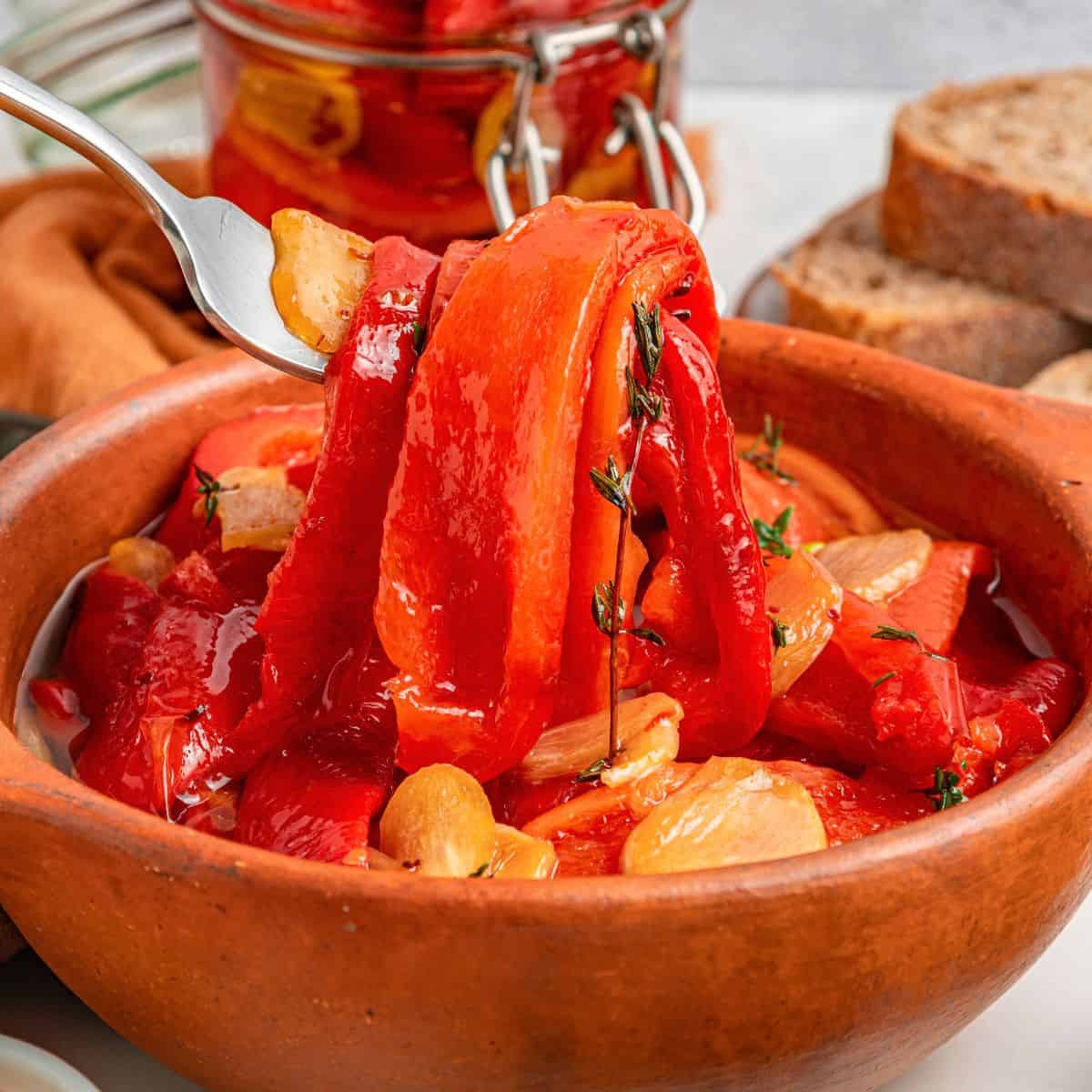 Roasted red peppers and garlic cloves in a brown bowl, garnished with herbs. A fork lifts some roasted red peppers. Sliced bread and a jar of peppers are in the background.