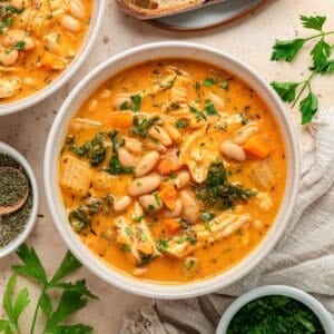 A bowl of creamy Chicken and White Bean Soup with tender chicken, carrots, greens, and herbs, garnished with parsley. Slices of bread and fresh herbs are arranged nearby on a light surface.