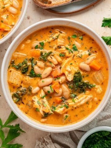 A bowl of creamy Chicken and White Bean Soup with tender chicken, carrots, greens, and herbs, garnished with parsley. Slices of bread and fresh herbs are arranged nearby on a light surface.