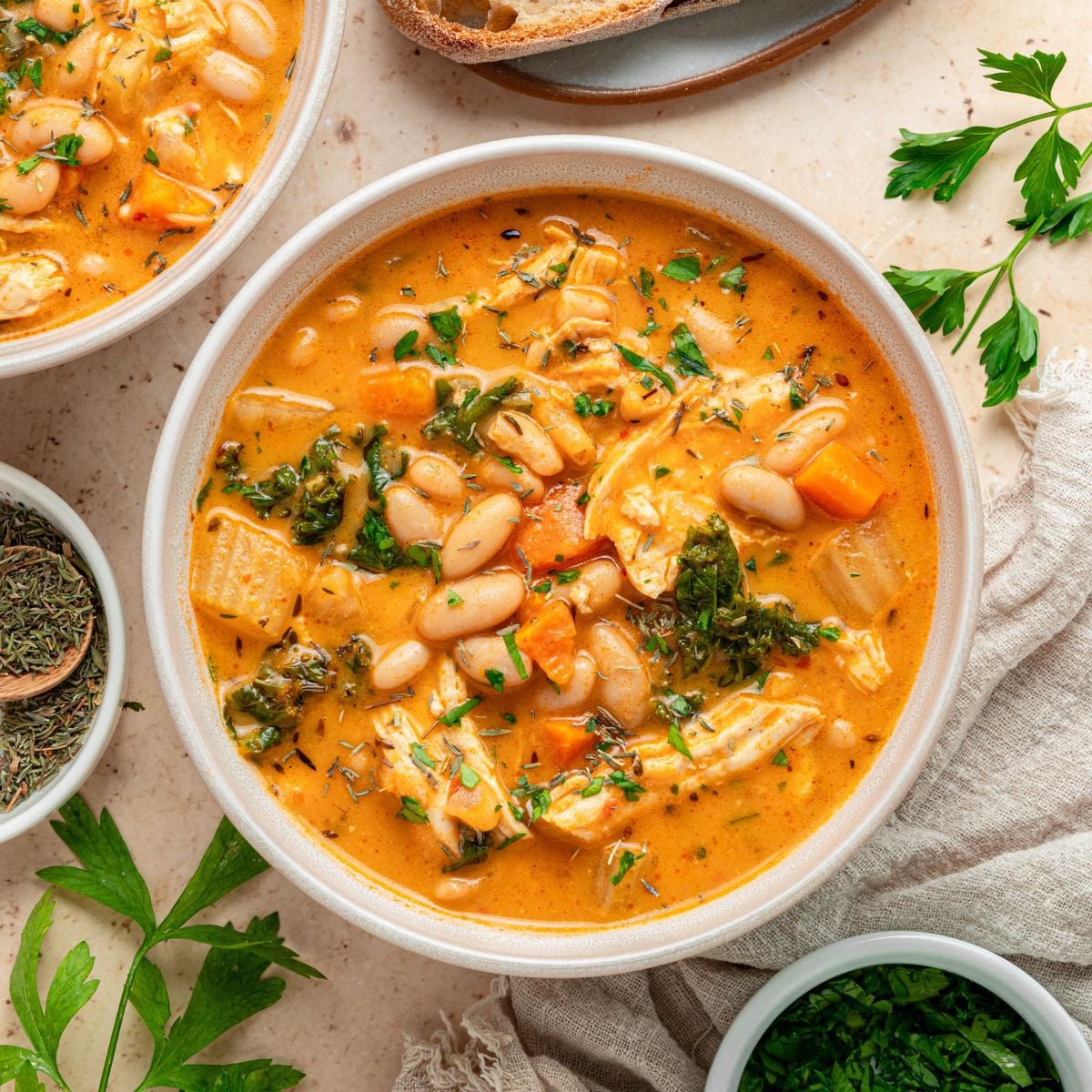 A bowl of creamy Chicken and White Bean Soup with tender chicken, carrots, greens, and herbs, garnished with parsley. Slices of bread and fresh herbs are arranged nearby on a light surface.