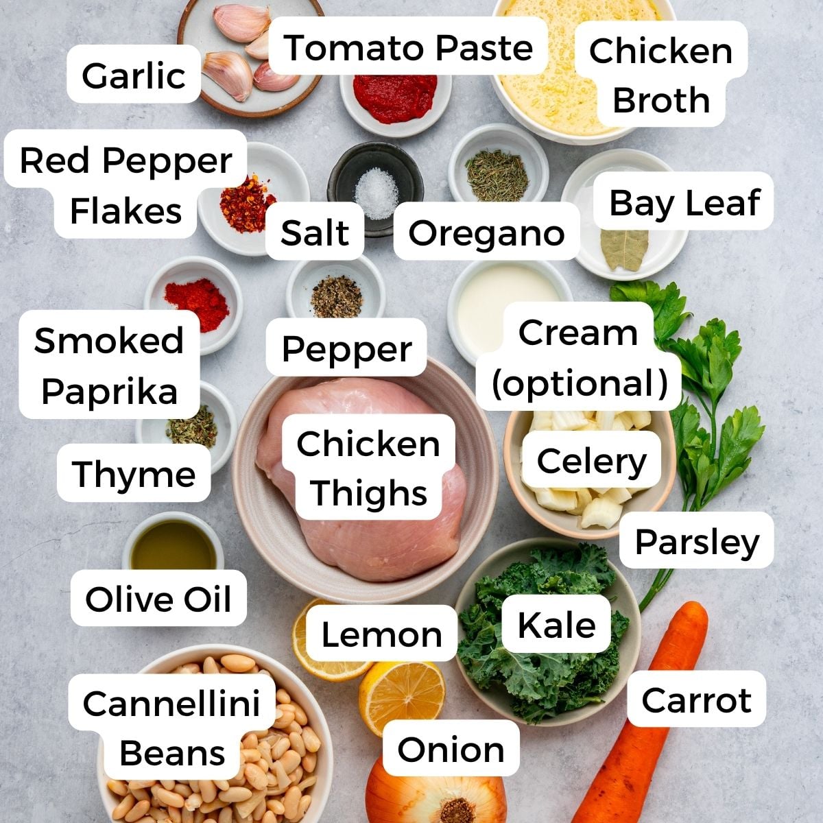 Top-down view of labeled ingredients for a chicken and white bean soup recipe, including chicken thighs, veggies, herbs, spices, beans, cream (optional), tomato paste, chicken broth, and seasonings arranged in white bowls and on a white surface.