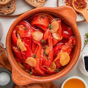 A clay bowl filled with roasted red peppers, sliced garlic, and herbs in oil sits at the center, surrounded by bread slices, honey, balsamic vinegar, chili flakes, and a salt dish.