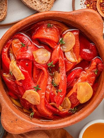 A clay bowl filled with roasted red peppers, sliced garlic, and herbs in oil sits at the center, surrounded by bread slices, honey, balsamic vinegar, chili flakes, and a salt dish.