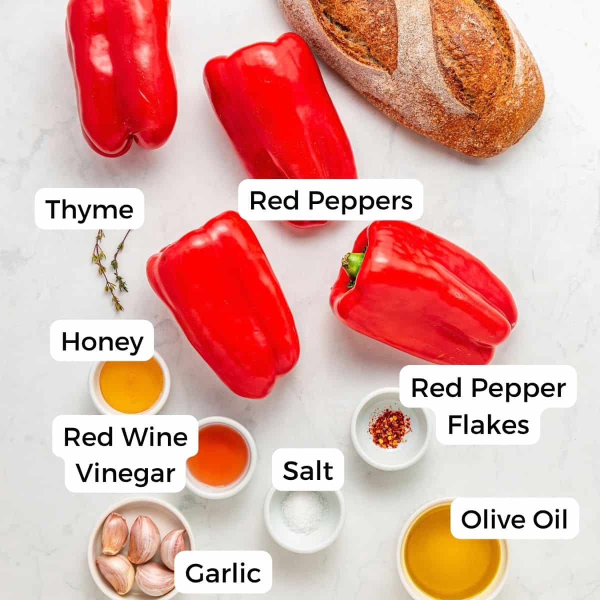 Ingredients on a white surface: roasted red peppers, a loaf of bread, garlic, thyme, honey, red wine vinegar, salt, red pepper flakes, and olive oil, with each item labeled.