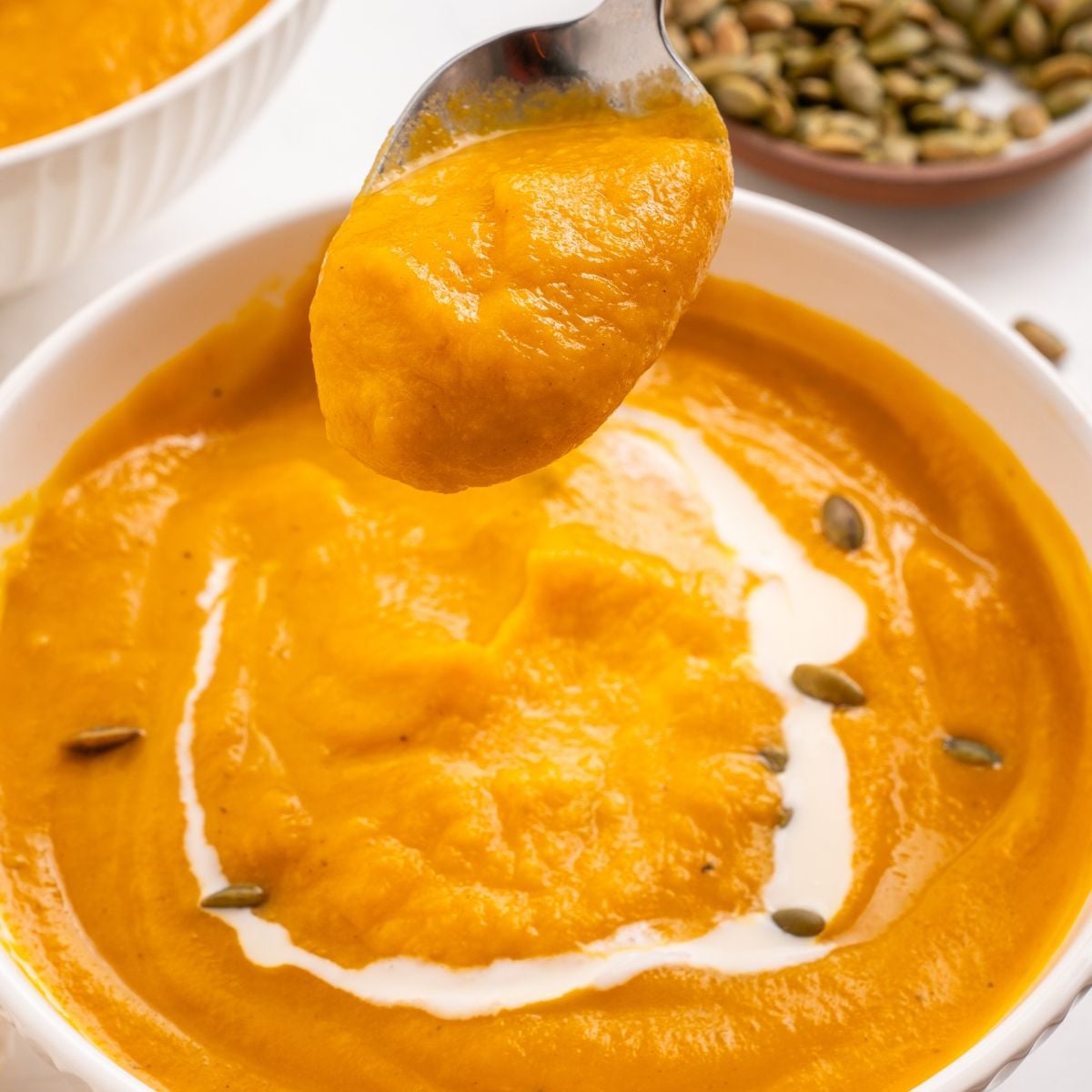 A spoonful of spiced carrot soup held above a bowl topped with a swirl of cream and scattered pumpkin seeds.