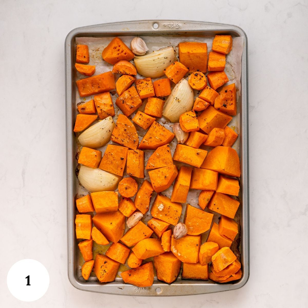 A metal baking sheet filled with roasted butternut squash cubes, onion wedges, and garlic cloves, prepped for a spiced butternut squash soup.