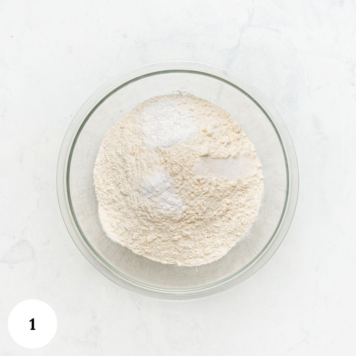 A glass bowl containing flour and piles of leavening ingredients for lemon ricotta cookies.