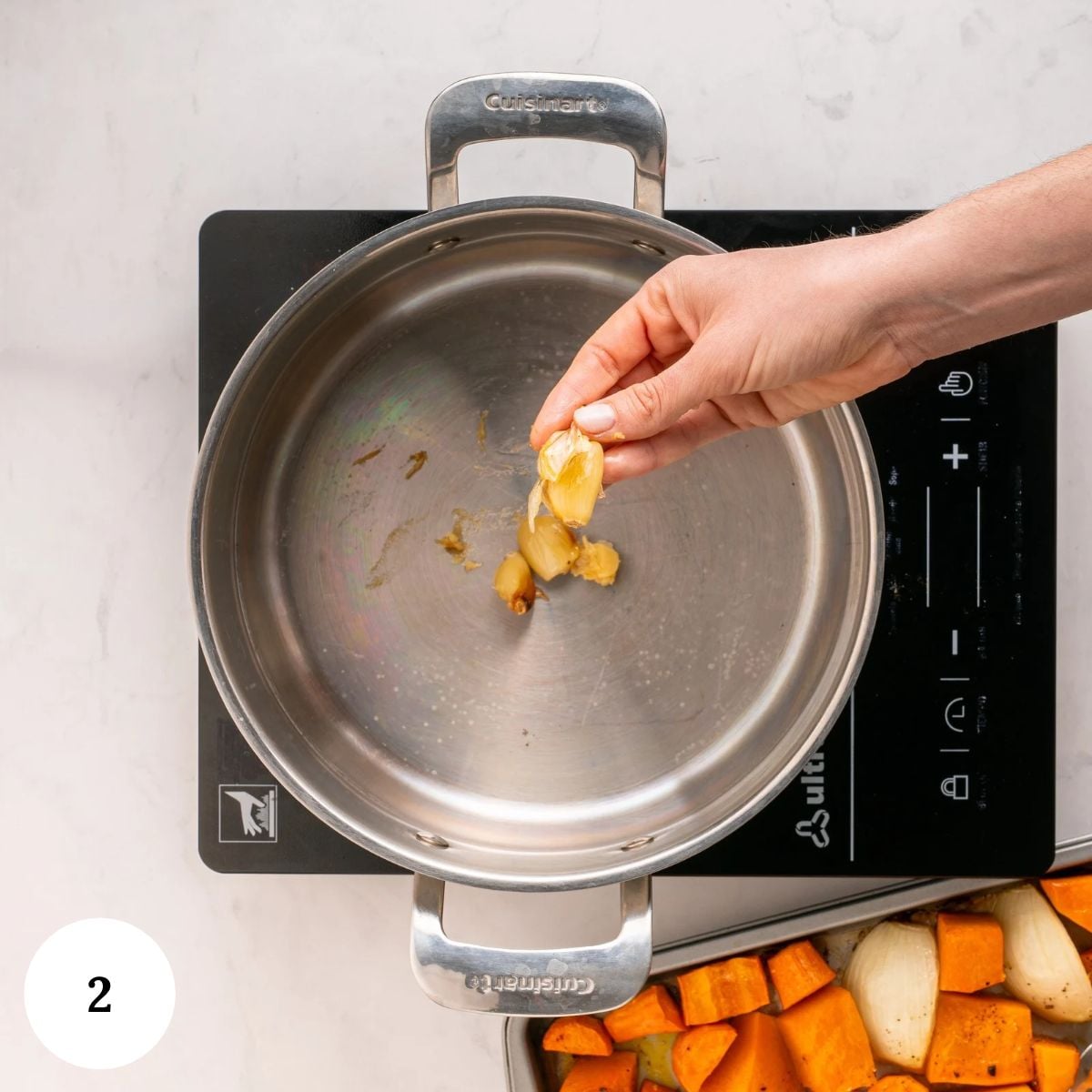A hand places a roasted garlic clove into a stainless steel pan on an induction cooktop to begin preparing a spiced carrot soup.