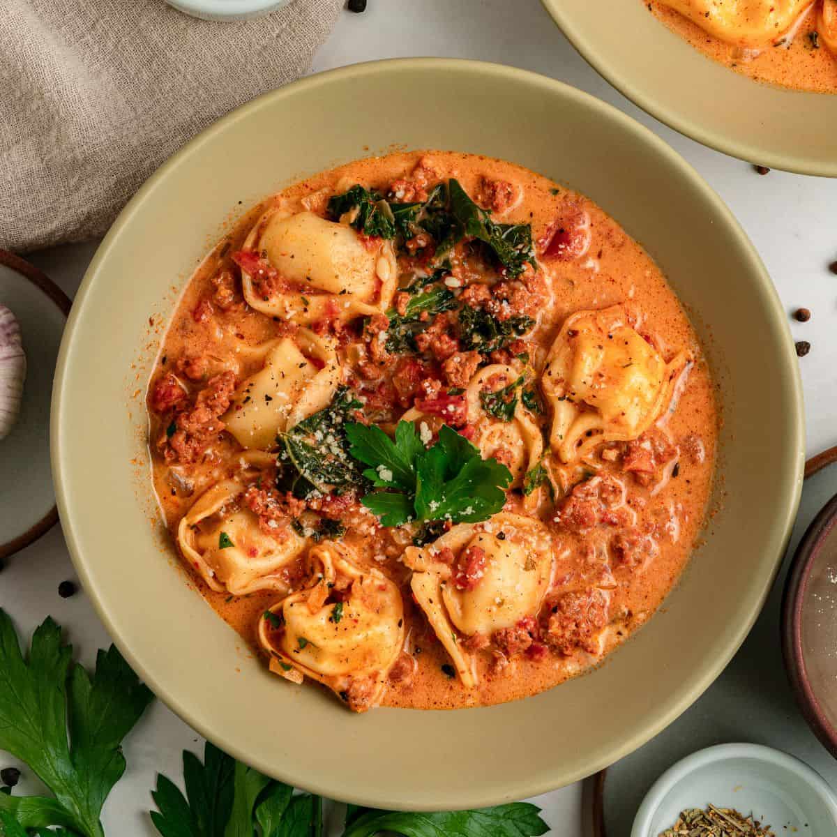 A bowl of creamy Tuscan tortellini soup with sausage, kale, and a garnish of fresh parsley.
