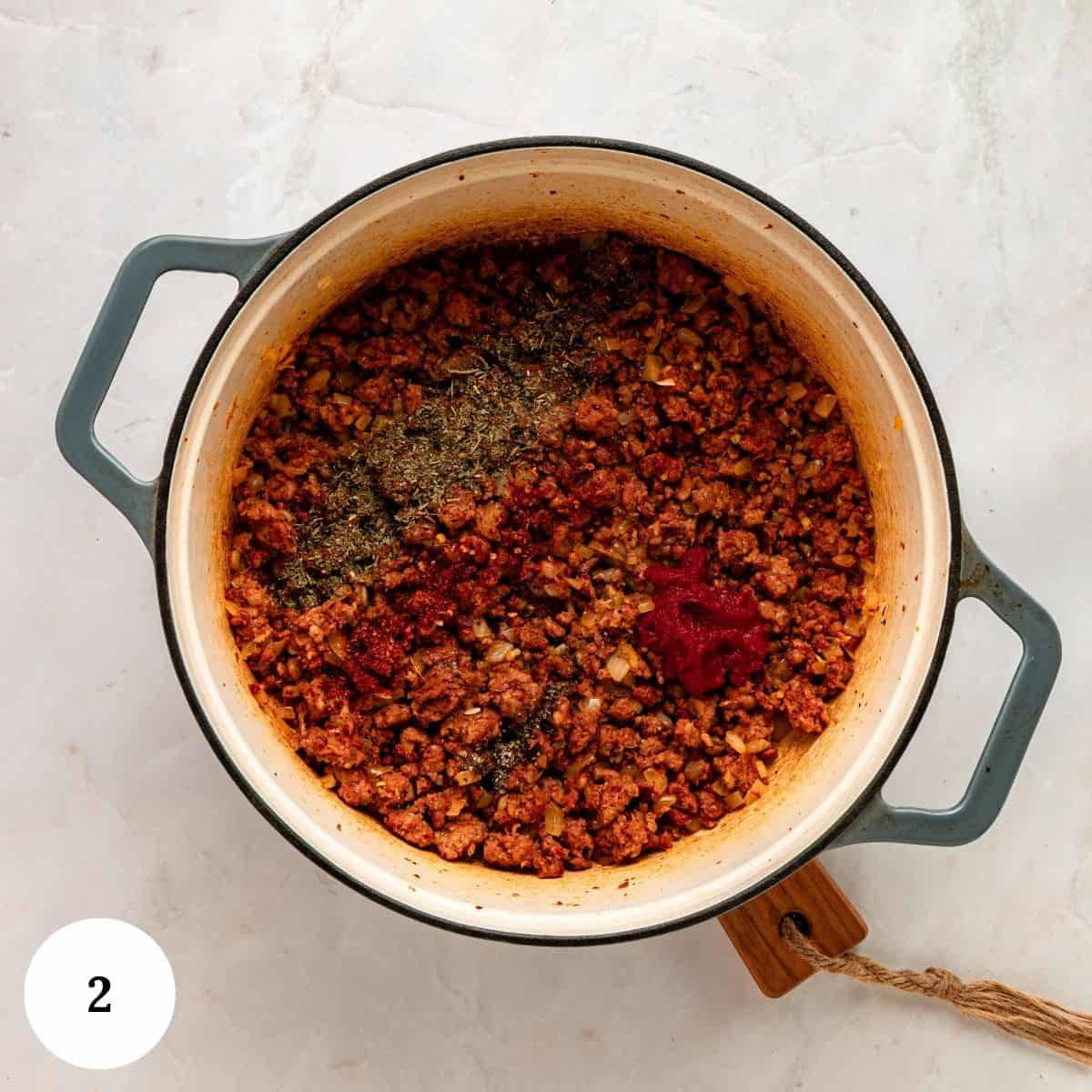 A Dutch oven containing browned ground meat, diced onions, a mound of dried herbs, and a dollop of tomato paste for Tuscan Tortellini soup.