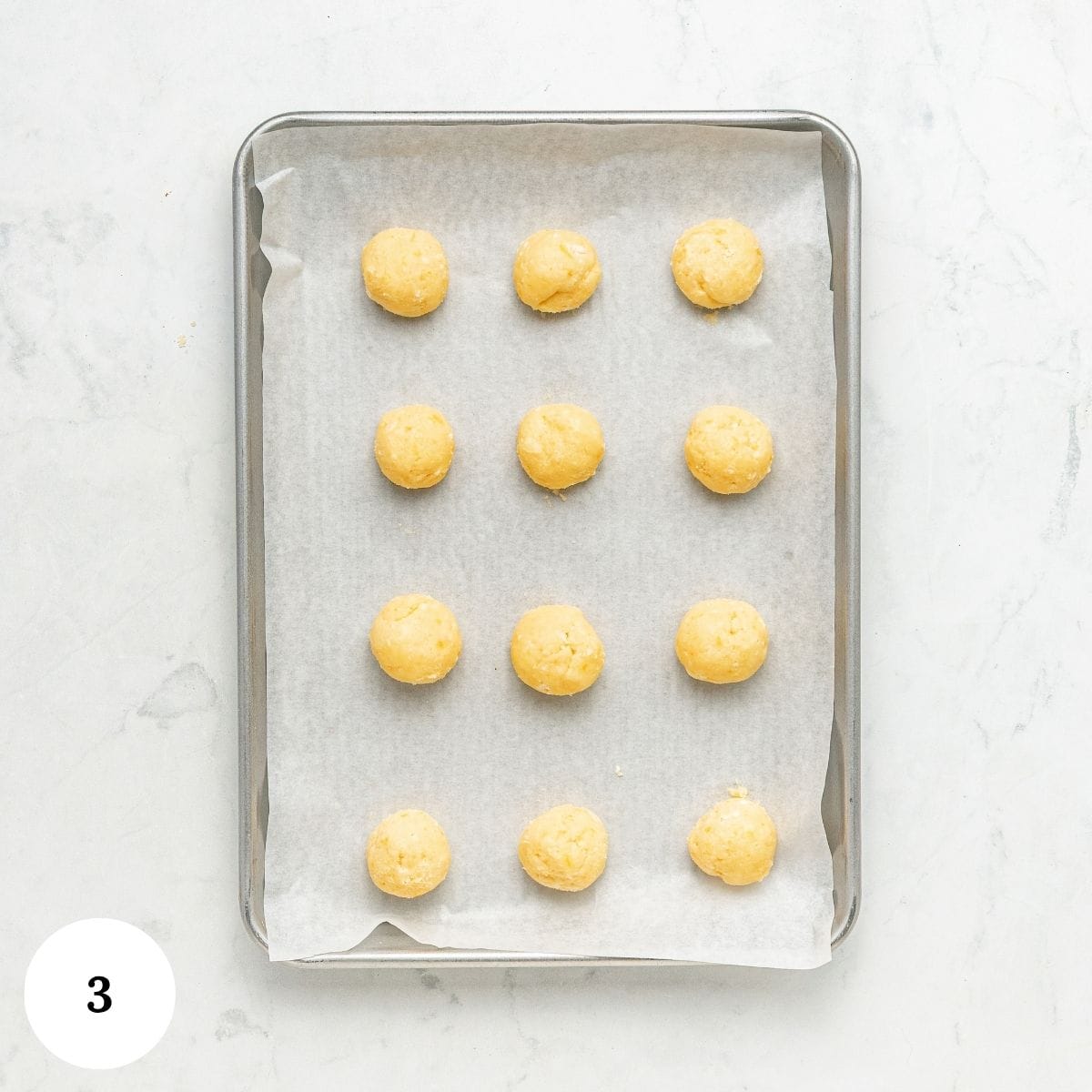Twelve balls of raw lemon ricotta cookie dough arranged in a grid on a parchment-lined baking sheet.