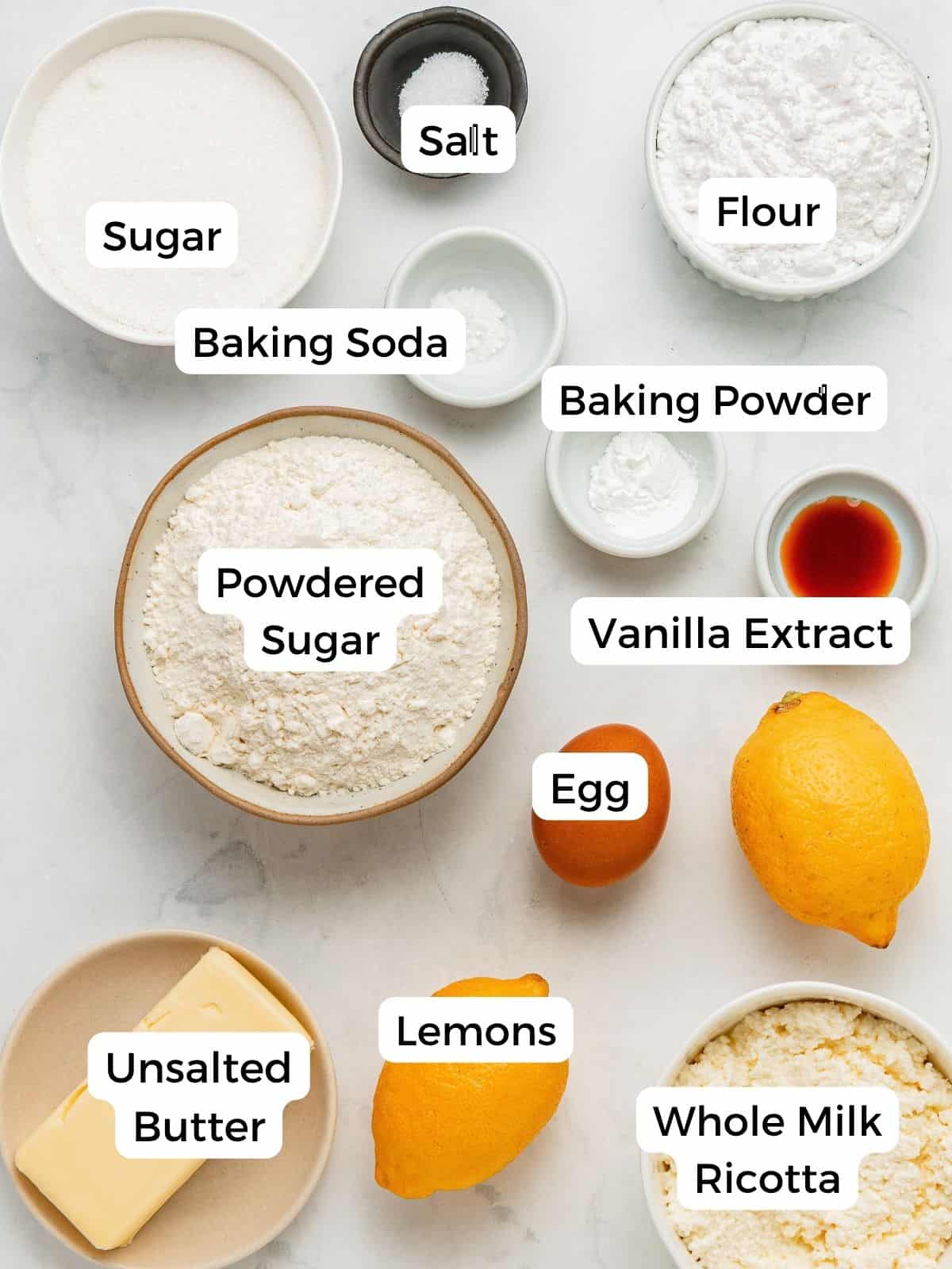Ingredients for lemon ricotta cookies arranged on a white surface, including sugar, salt, flour, baking soda, baking powder, vanilla extract, an egg, lemons, butter, and ricotta, each labeled with its name.