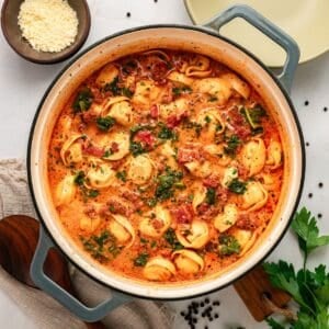 Tuscan Tortellini soup with ground meat and kale in a creamy red tomato-based sauce, served in a Dutch oven with a side of grated cheese.